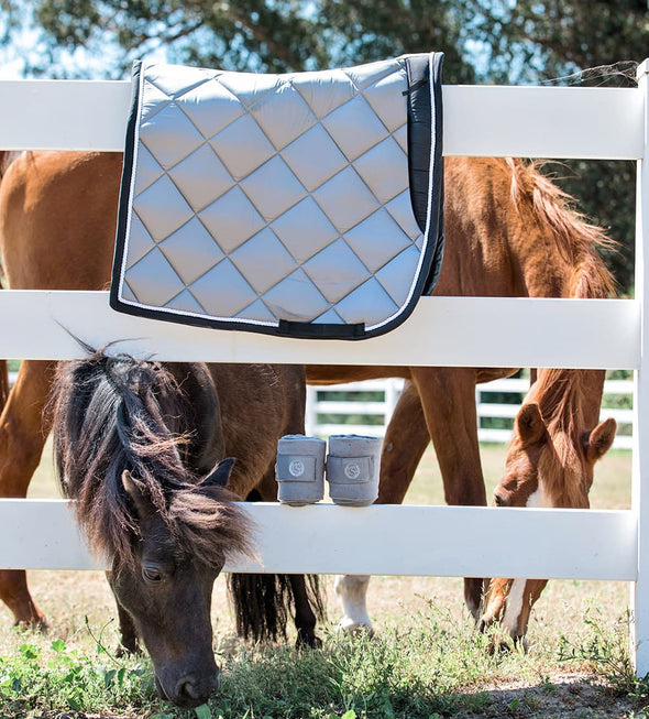 Dressage saddle pad and polo wraps on a white vinyl fence. Two horses are in the background with one peaking it's head through the fence. Gray Dressage Bundle with Fly Veil, Polo Wraps and a Dressage saddle pad. Gray in color with semi shiny material.